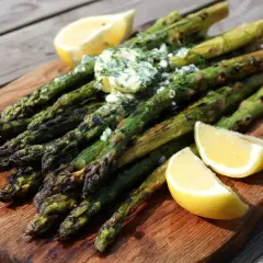 Grilla sparris med smör och dill, serverad med citronklyftor på en träbricka. Perfekt sommarbuffé!