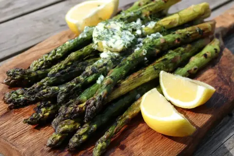 Grilla sparris med smör och dill, serverad med citronklyftor på en träbricka. Perfekt sommarbuffé!