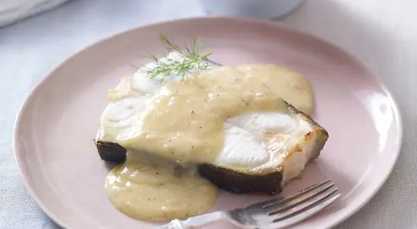 En bit fisk täckt med krämig beurre blanc-sås, garnerad med dill, serveras på en rosa tallrik med en gaffel.