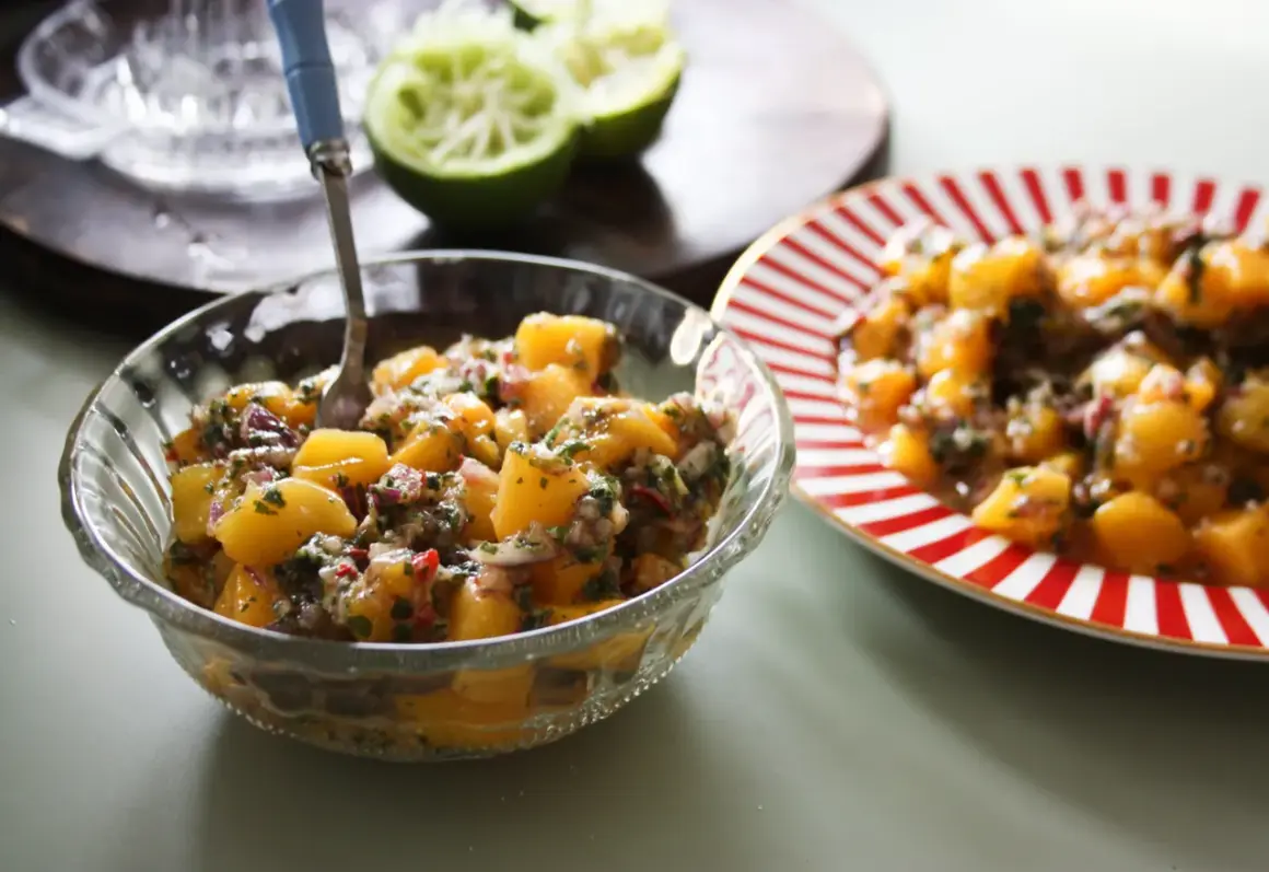 En skål med färgglad mangosalsa, med lime och en röd-vit randig tallrik i bakgrunden.