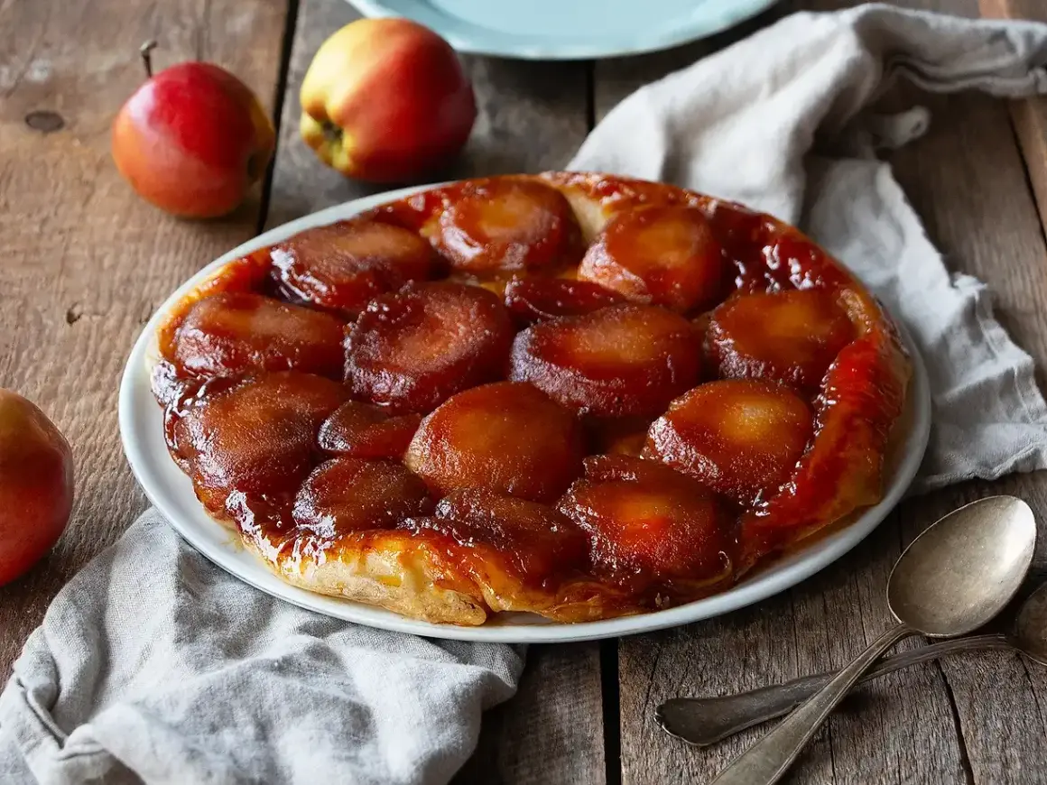 En gyllene tarte tatin med karamelliserade äpplen på en vit tallrik, omgiven av äpplen och en linneduk.