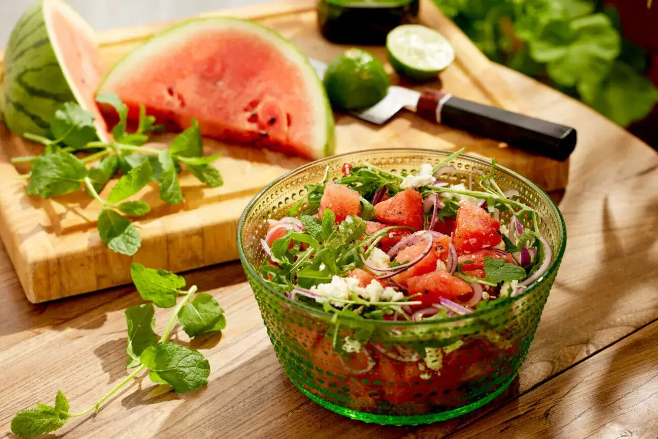 En sk&aring;l med godaste melonsalladen, fylld med vattenmelon, fetaost, r&ouml;dl&ouml;k och mynta. Bredvid ligger en halv vattenmelon och lime p&aring; en sk&auml;rbr&auml;da.