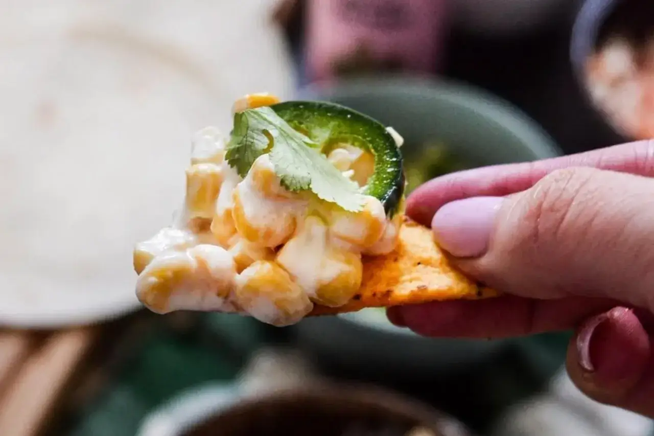 En hand h&aring;ller en nachochips med kr&auml;mig majs, jalape&ntilde;o och koriander.