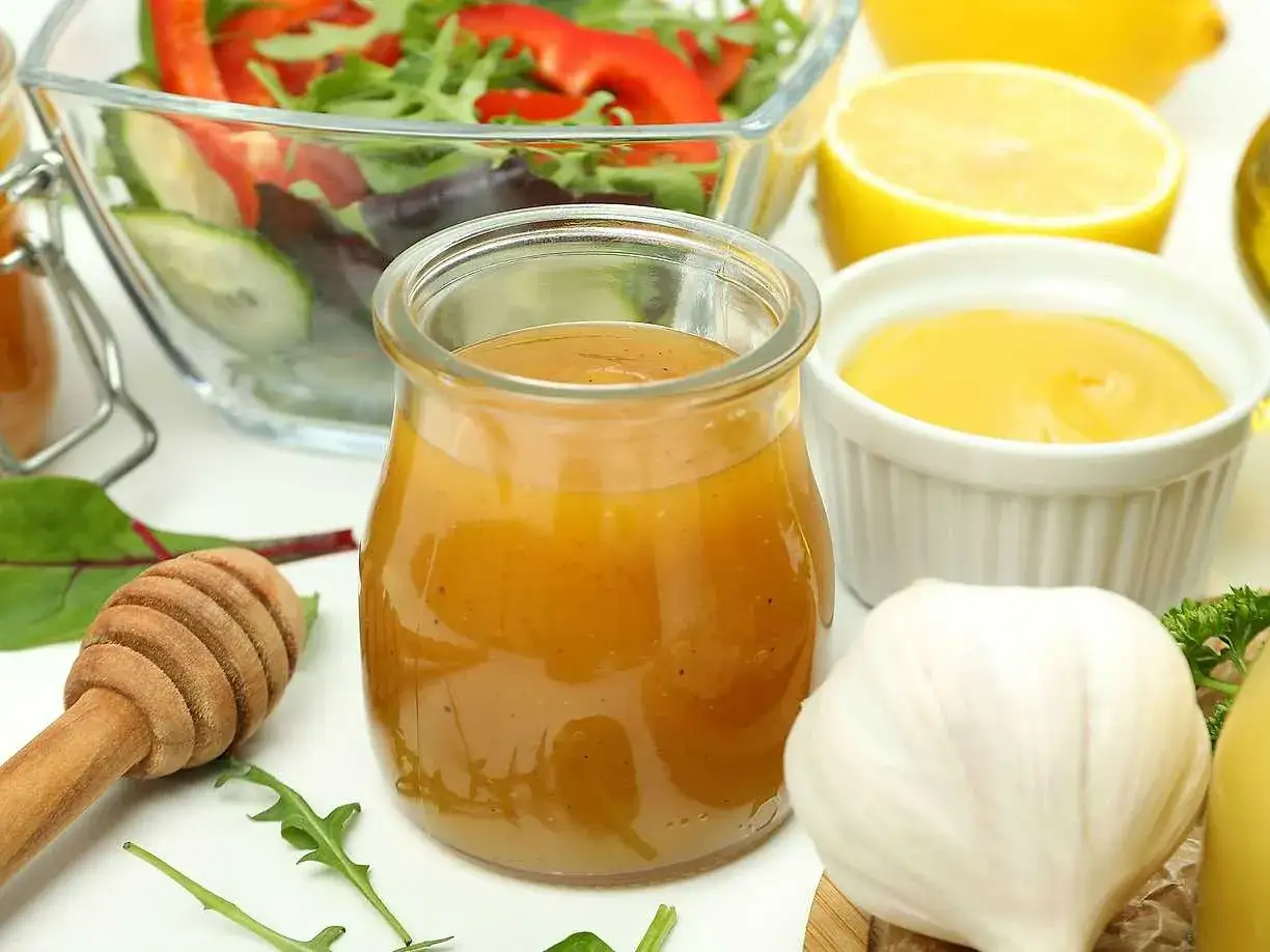 En burk med gyllene dressing, perfekt som godaste dressingen till sallad. Ingredienser som citron, vitlök och honung syns bredvid.