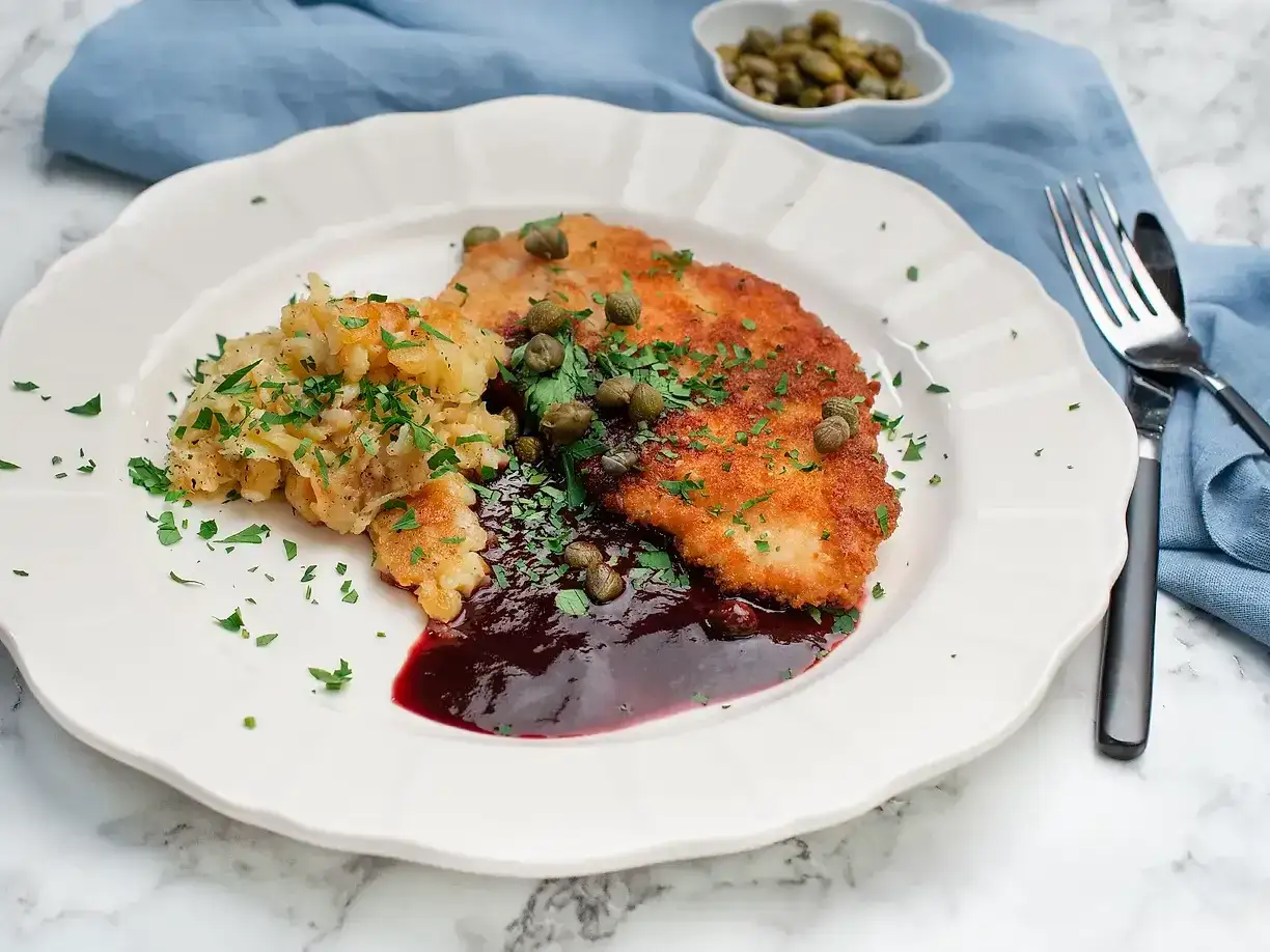 En tallrik med schnitzel, potatis och en m&ouml;rk s&aring;s till schnitzel, garnerad med kapris och persilja.