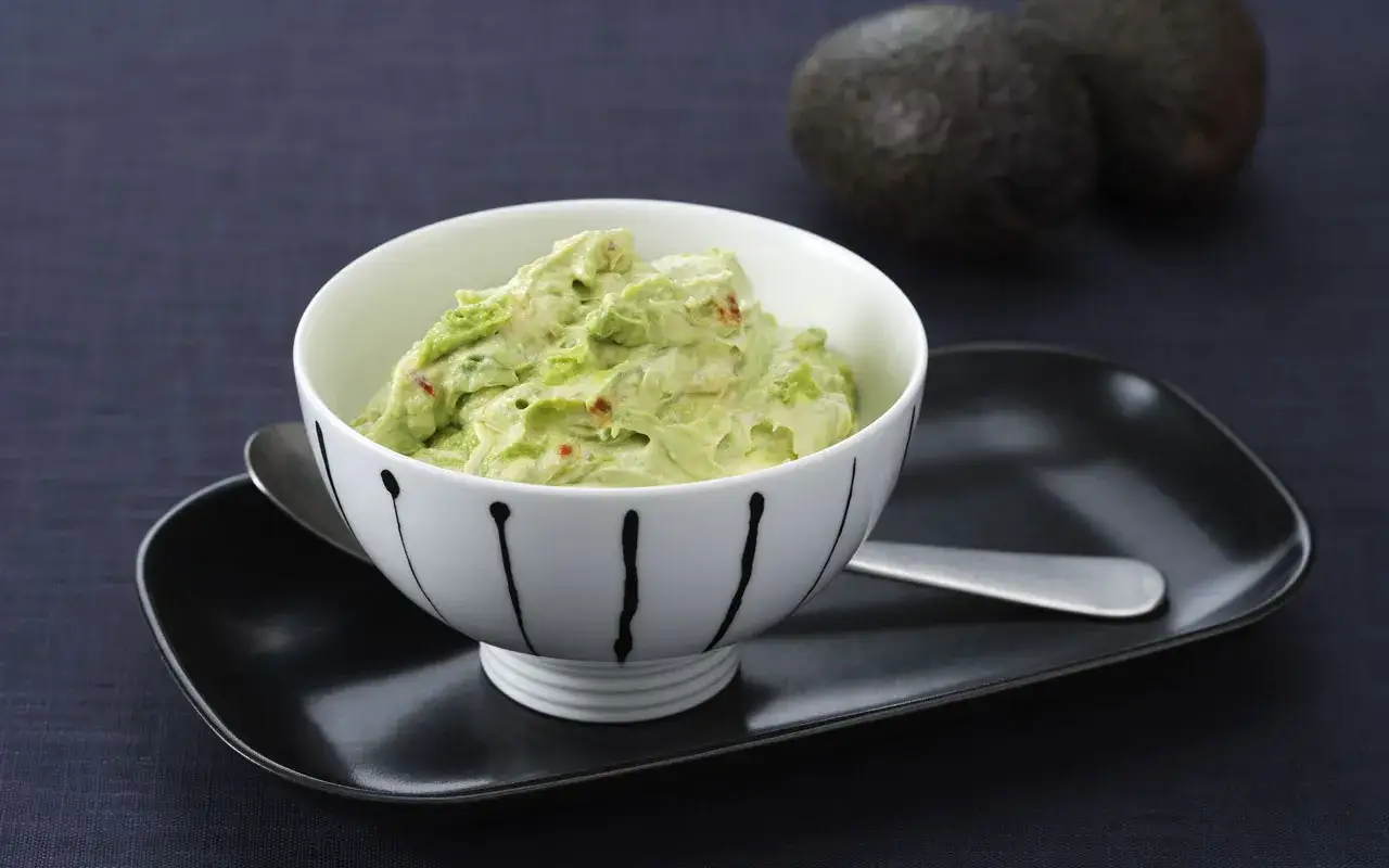 Krämig guacamole med en touch av crème fraîche, serverad i en vacker skål. Två avokados syns i bakgrunden.