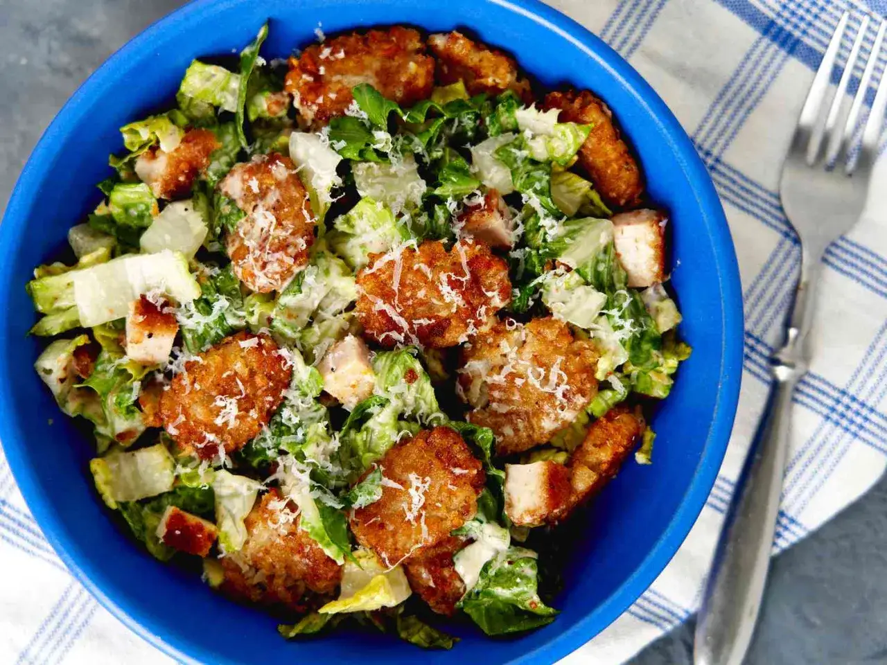 En blå skål med caesarsallad, kycklingbitar och krispiga tater tots, toppad med riven parmesan.