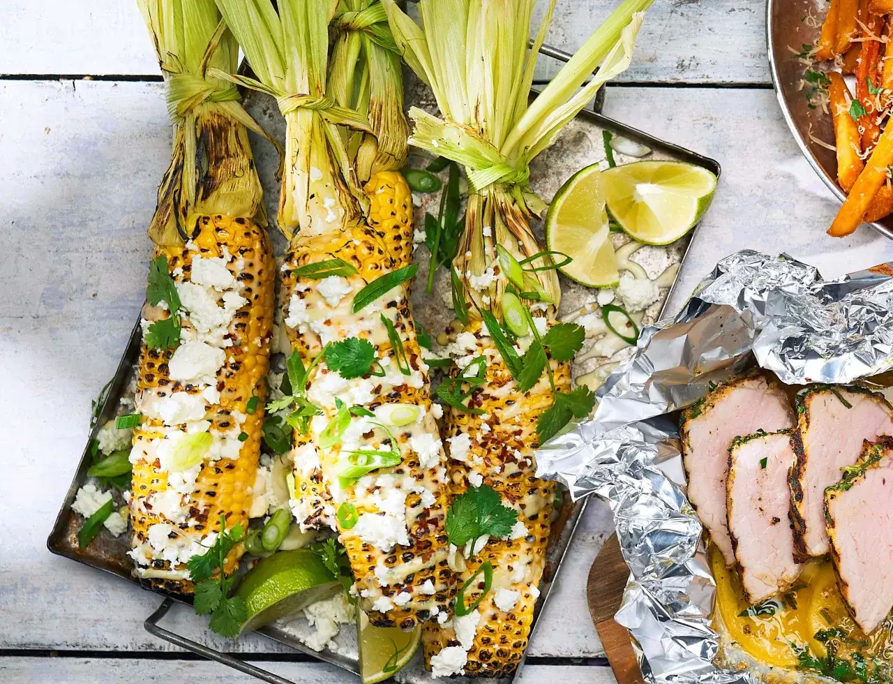 Grillade majskolvar, corn ribs, toppade med ost, &ouml;rter och lime. Bredvid ligger skivat fl&auml;skk&ouml;tt i folie.